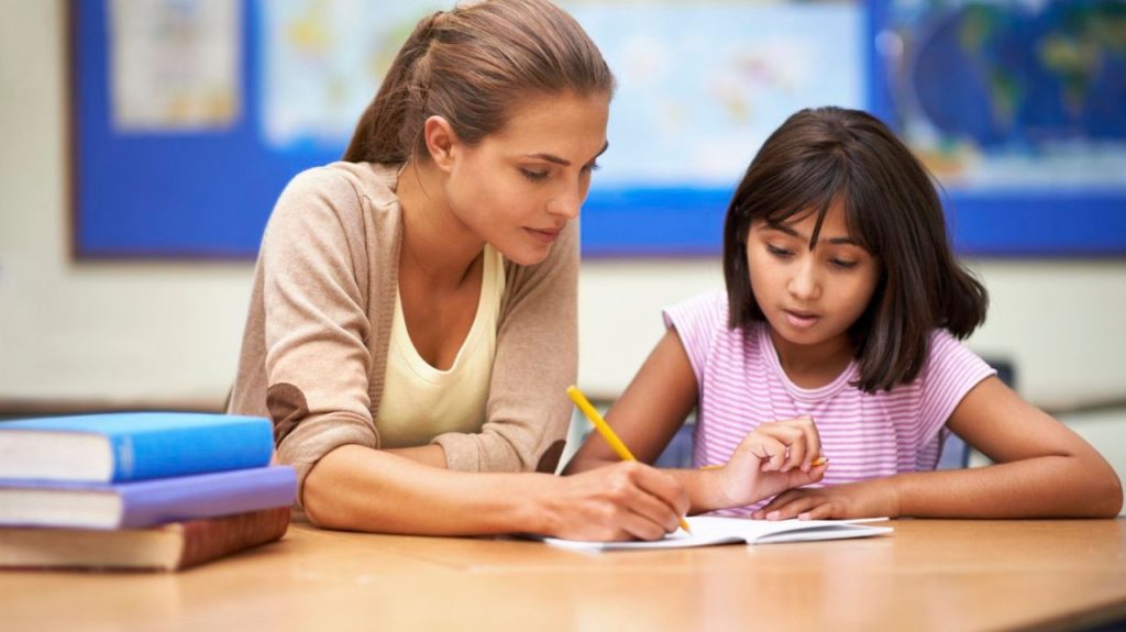 Young female student studying with a teacher in a classroom. They are working on some homework and have books on the desk